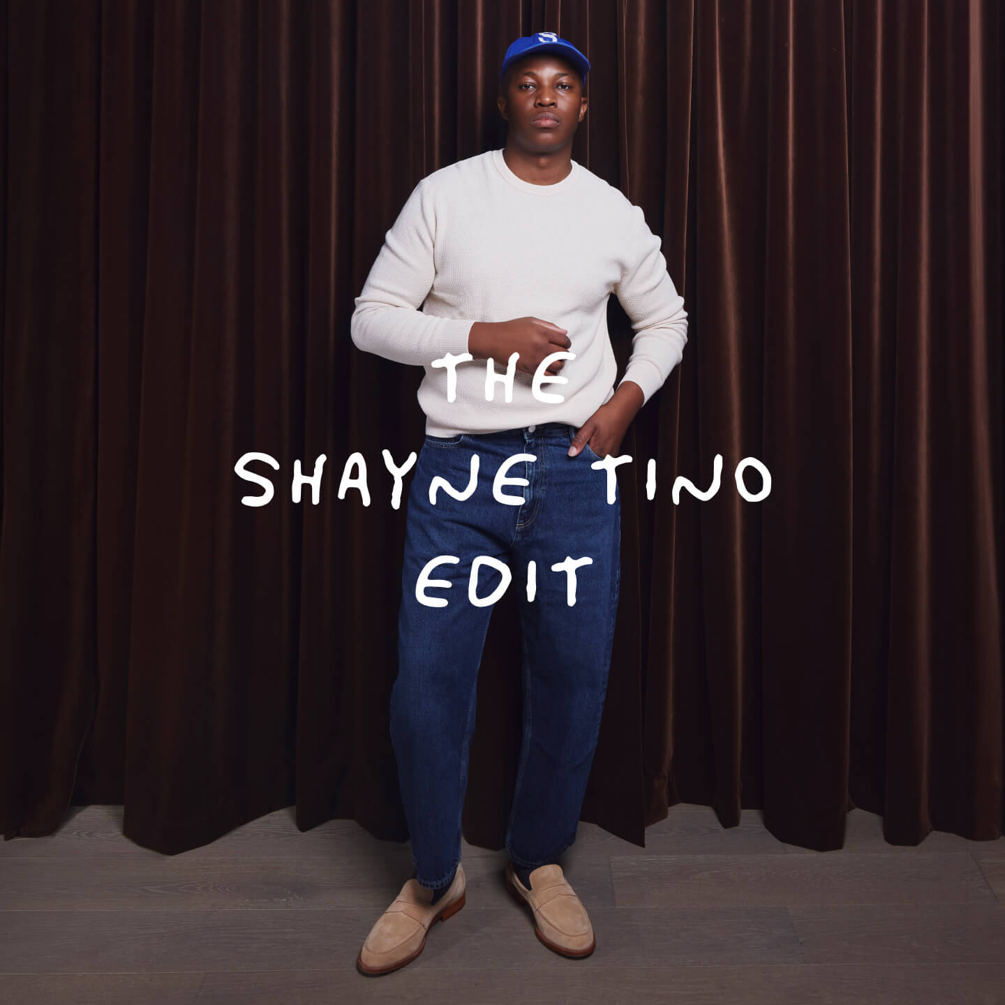 Man wearing a white sweater and blue jeans with text 'The Shayne Tino Edit' against a brown curtain background.