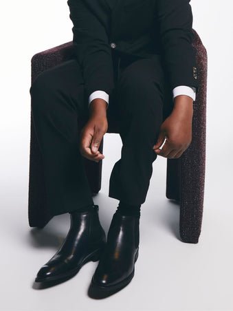 Person wearing a black suit and shoes sitting on a chair with a white background