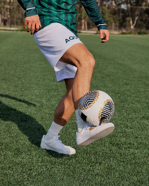 Person kicking a soccer ball on a grass field, wearing white shorts with 'Aquila' branding.